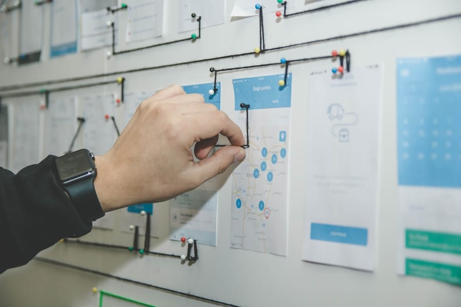 A person adjusts black string connecting pins on a whiteboard covered with papers, diagrams, and blueprints, possibly planning a project or mapping a process. The person is wearing a smartwatch.