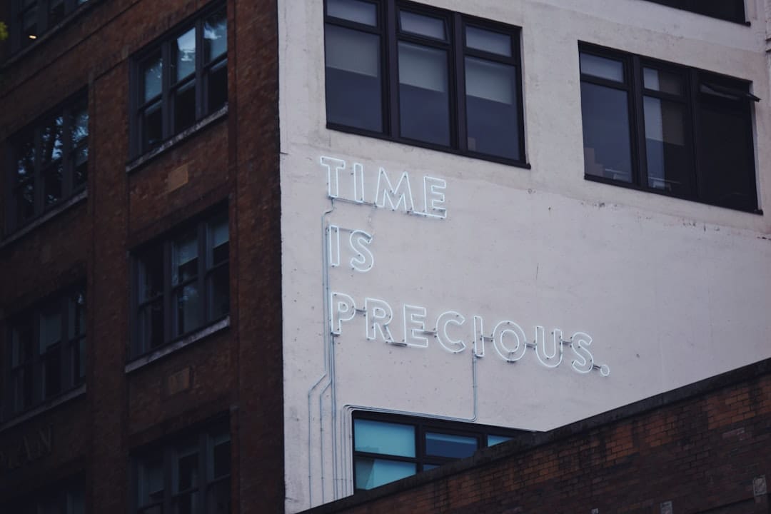 A building wall displays a white neon sign reading “TIME IS PRECIOUS.” The surrounding architecture features brick and large windows.