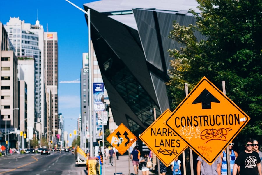 Busy city street with modern buildings, trees, and multiple orange construction signs, including one reading “CONSTRUCTION” with graffiti, and people walking on the sidewalk under a clear blue sky.