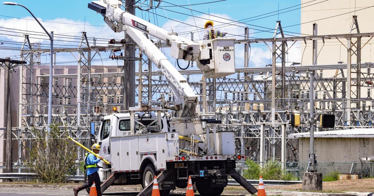 Two utility workers in safety gear operate a bucket truck near power lines at an electrical substation, with orange cones placed around the vehicle for safety. One worker stands on the ground, while the other works in the raised bucket.