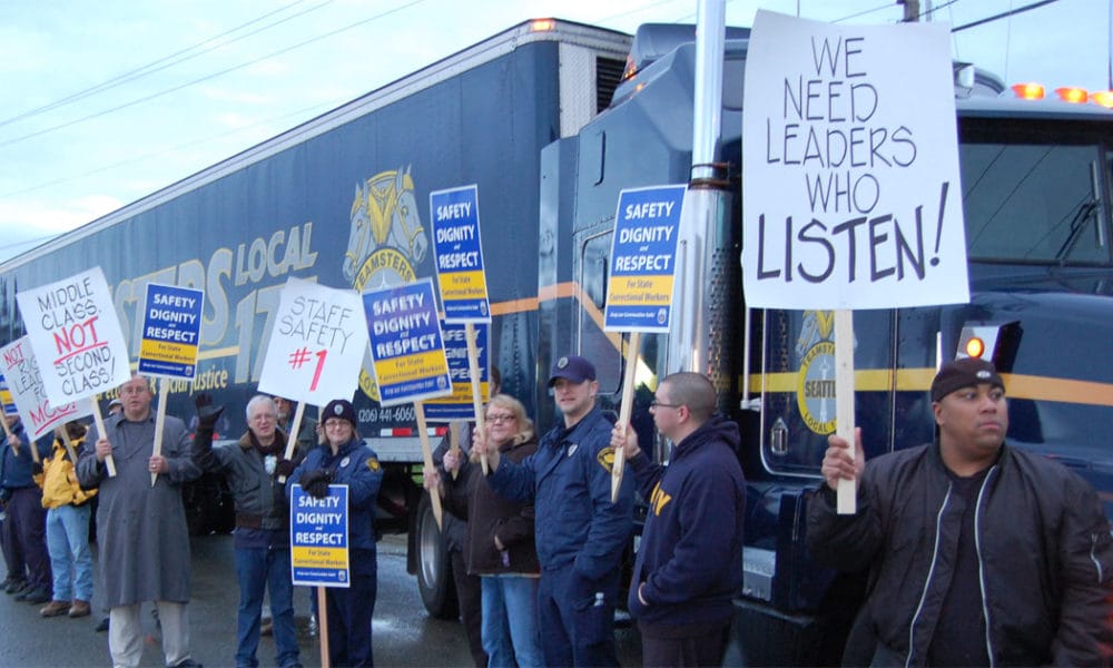 A group of people stand in front of a large truck, holding signs with messages such as “SAFETY DIGNITY RESPECT,” “STAFF SAFETY #1,” and “WE NEED LEADERS WHO LISTEN!” during a protest or demonstration.