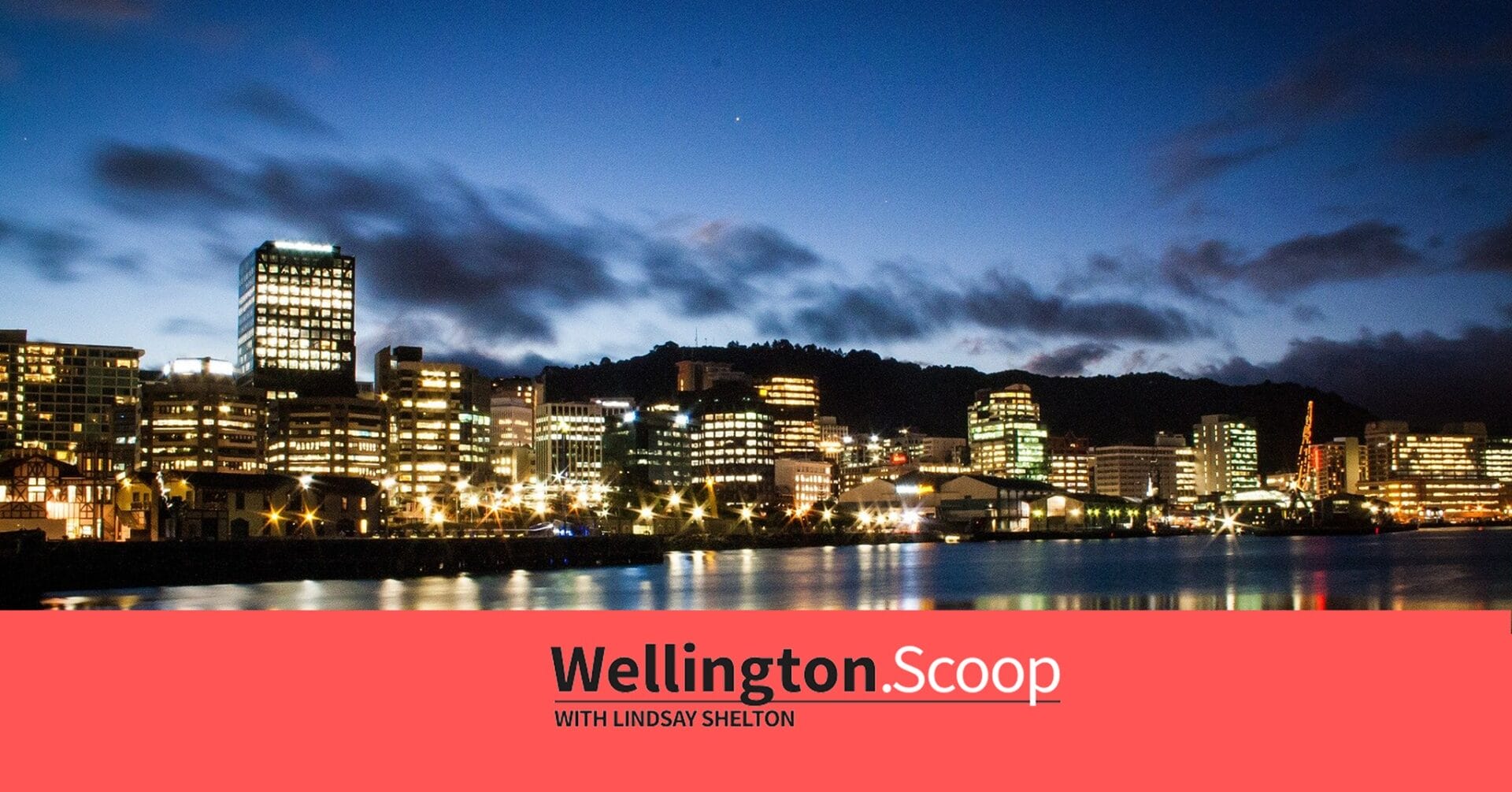 A view of Wellington city’s skyline at dusk, with illuminated buildings reflecting on the waterfront. The sky is deep blue with scattered clouds. A red banner at the bottom reads “Wellington Scoop with Lindsay Shelton.”.