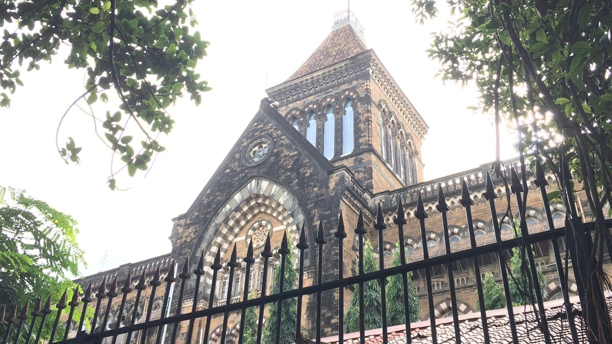 A historic stone building with arched windows and a clock tower, partially obscured by a black metal fence and framed by tree branches, seen in bright daylight.