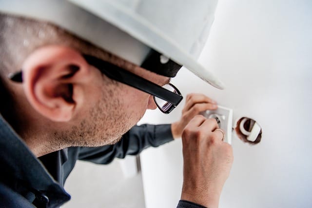 A person wearing glasses and a white hard hat uses a screwdriver to install or repair a wall electrical outlet, demonstrating the importance of Electrical Safety during maintenance.