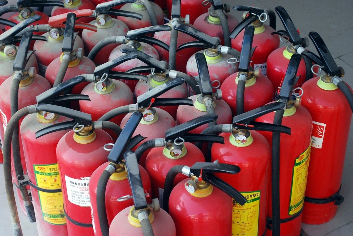 A large group of red fire extinguishers with black hoses and handles are clustered together on a light-colored floor. Labels and maintenance tags are visible on several of the extinguishers.