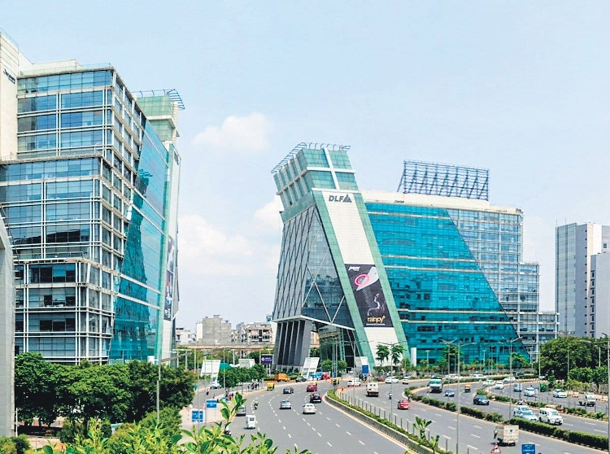 A modern cityscape with glass-covered office buildings, wide roads, green trees, and light traffic under a clear blue sky. The buildings have unique angular designs and prominent signage.