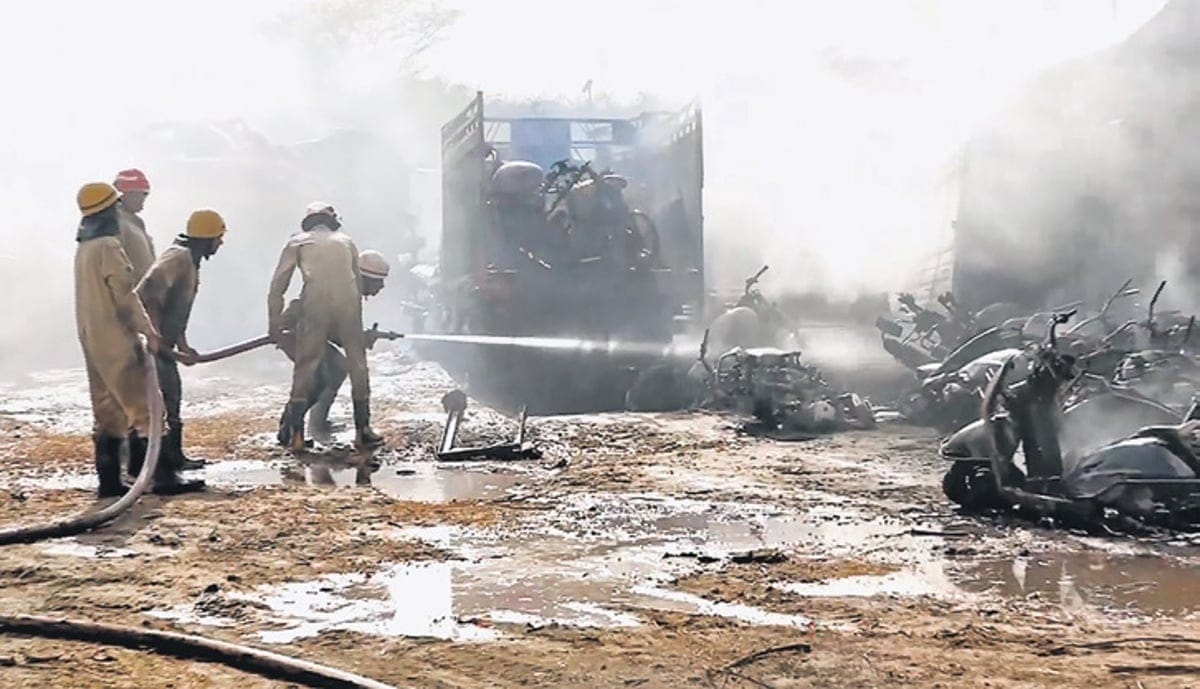 Firefighters in protective gear spray water toward a smoking fire at a scrapyard, with damaged vehicles and debris scattered on muddy ground under a hazy sky.