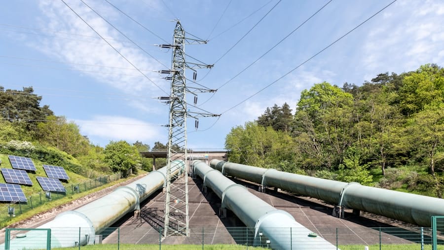 Large pipelines run up a grassy hill flanked by trees, with solar panels on the left and a metal electricity pylon in the center under a partly cloudy sky.
