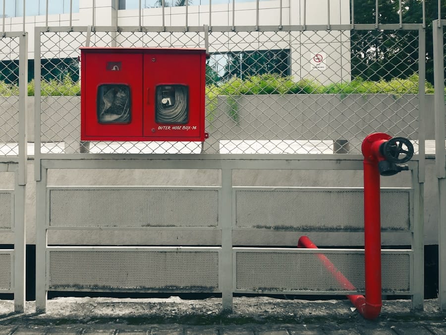 A red fire hose cabinet and a red pipe with a valve are mounted on a fence in front of a concrete wall. Grass and a modern building can be seen in the background.