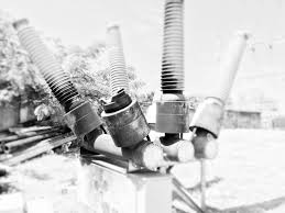 Black and white image of three large electrical insulators mounted outdoors, each with a ribbed cylindrical shape and attached to metal bases, essential components in power grid infrastructure, with blurred background of foliage and structures.