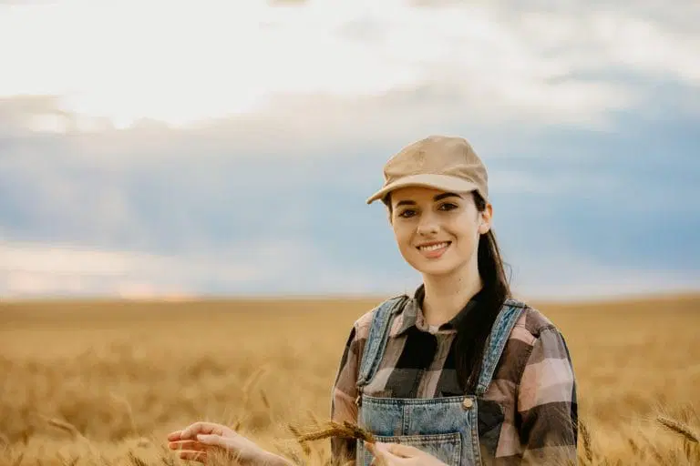 A young woman in overalls and a beige cap stands smiling in a golden wheat field under a partly cloudy sky, highlighting the importance of air quality analysis for healthy rural environments.