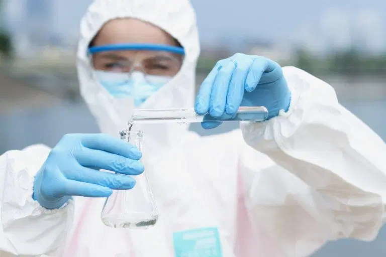 A scientist in full protective gear and blue gloves pours a clear liquid from a test tube into a conical flask outdoors during air quality analysis.