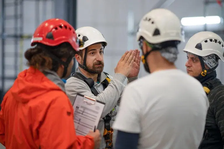 A man in a safety harness and helmet gestures while discussing a safety audit with three others in helmets and work gear; one person holds a clipboard.