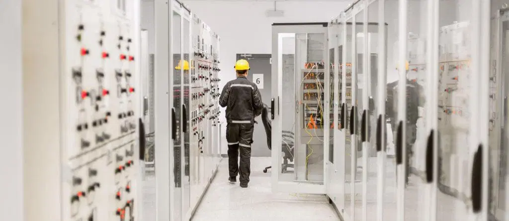 A worker in a yellow hard hat and protective clothing walks through a corridor lined with electrical control panels, performing Power Quality Analysis in an industrial facility.