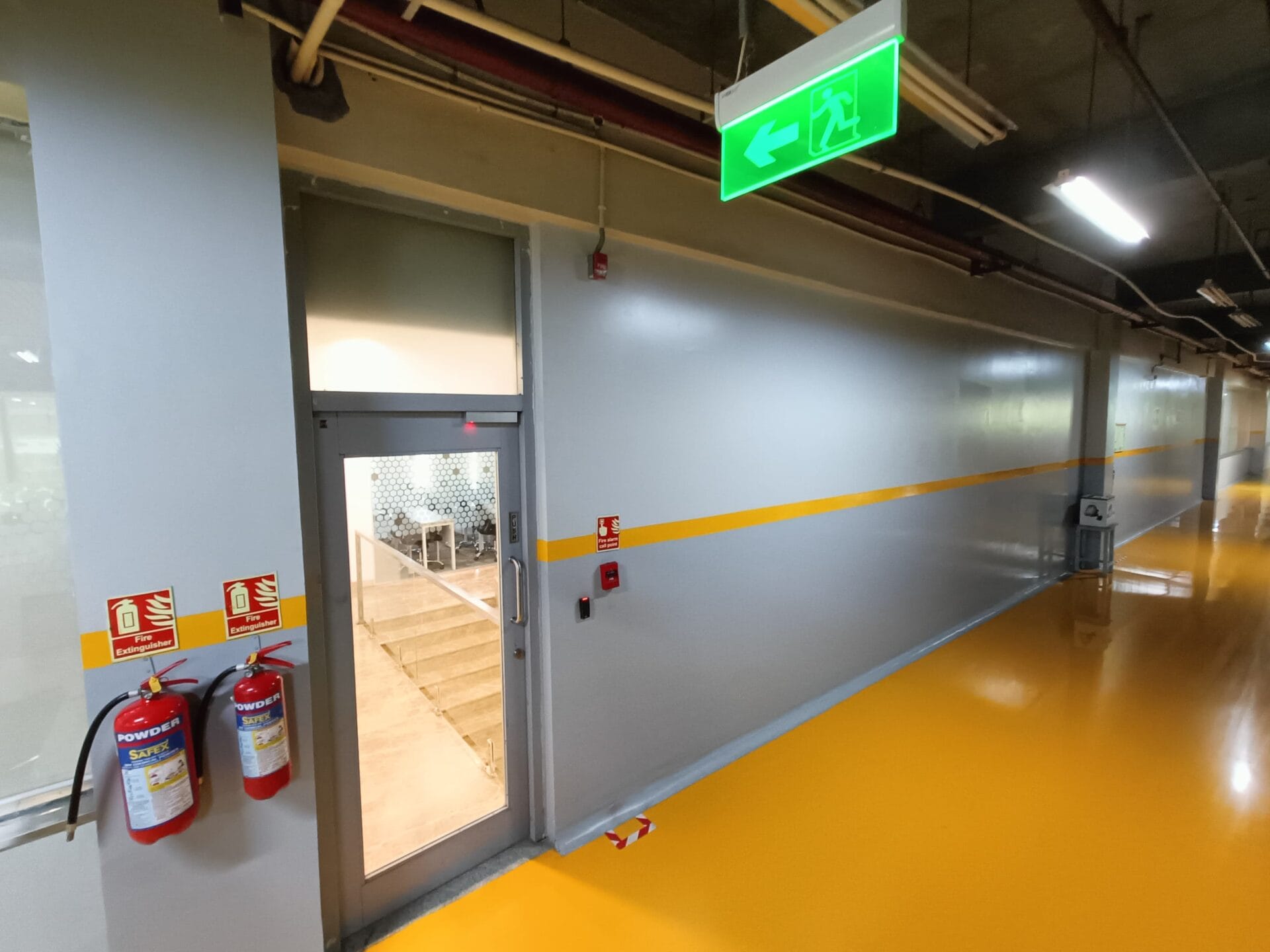 A hallway in Goa featuring a yellow floor stripe, gray walls, red fire extinguishers, and an emergency exit door under a green left-arrow sign—ideal for a Hotel Safety or Fire Safety audit by Elion Technologies.