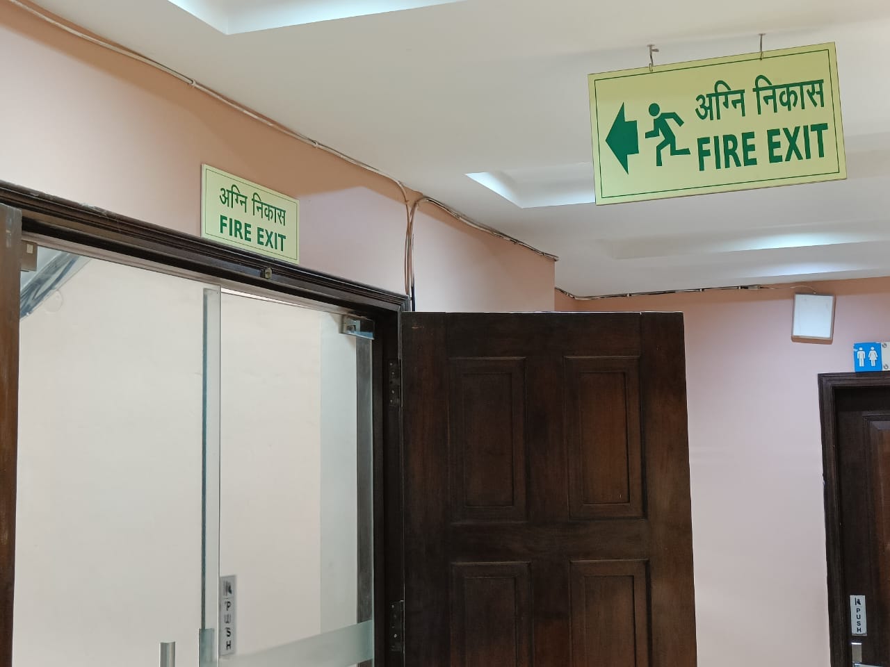 A hallway with brown doors and green Fire Exit signs in English and Hindi above a glass door, indicating the direction for emergency exit.