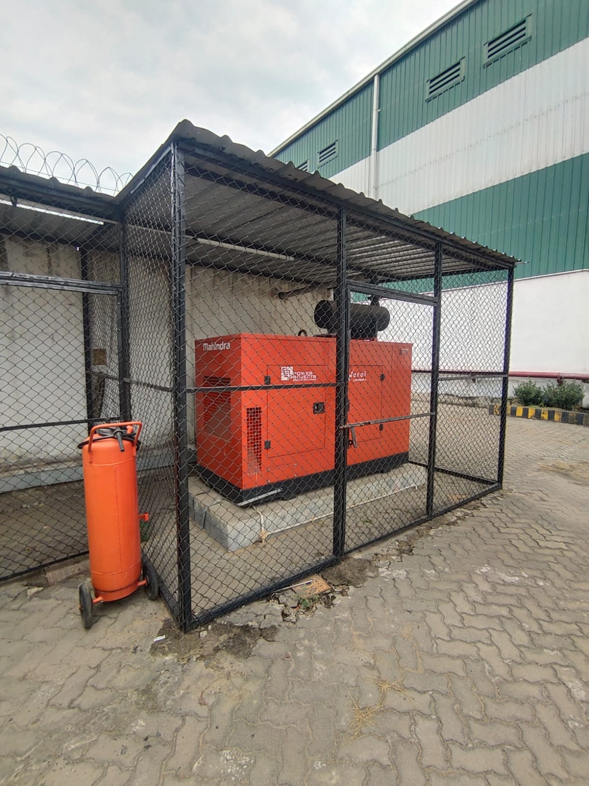 A large orange generator is enclosed in a black wire mesh cage with a corrugated metal roof, demonstrating effective energy safety measures. An orange fire extinguisher stands outside this setup beside a green and white industrial building.