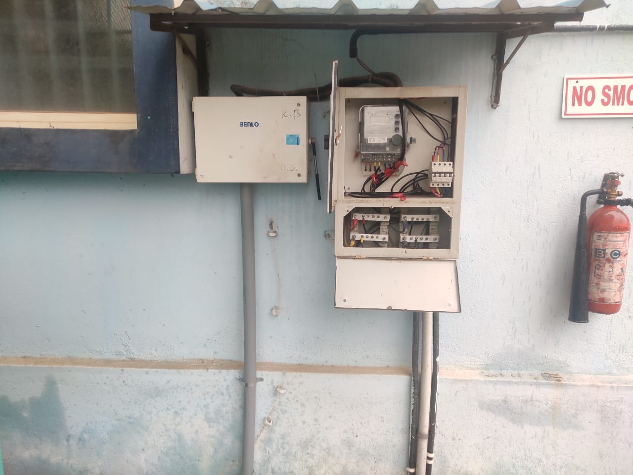 An open electrical control panel with visible wiring and meters is mounted on a light blue wall near a fire extinguisher and a “NO SM” sign, ready for an electrical safety audit. A gray box labeled “BEND” is mounted above the panel.