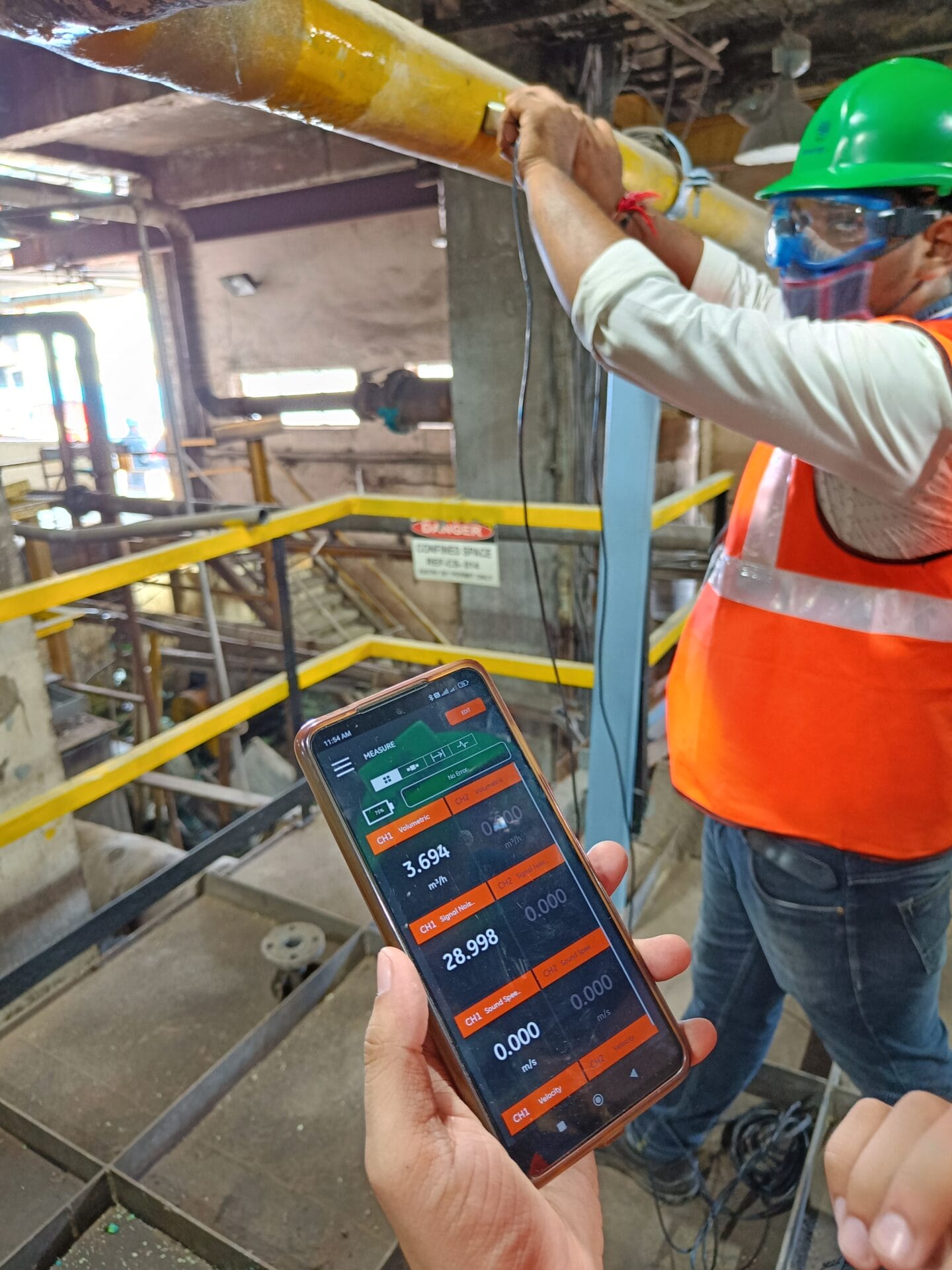 A person holds a smartphone displaying industrial data for Water Risk Assessment, while another worker in safety gear adjusts equipment on a yellow pipe in a factory. Safety railings and warning signs are visible in the background.