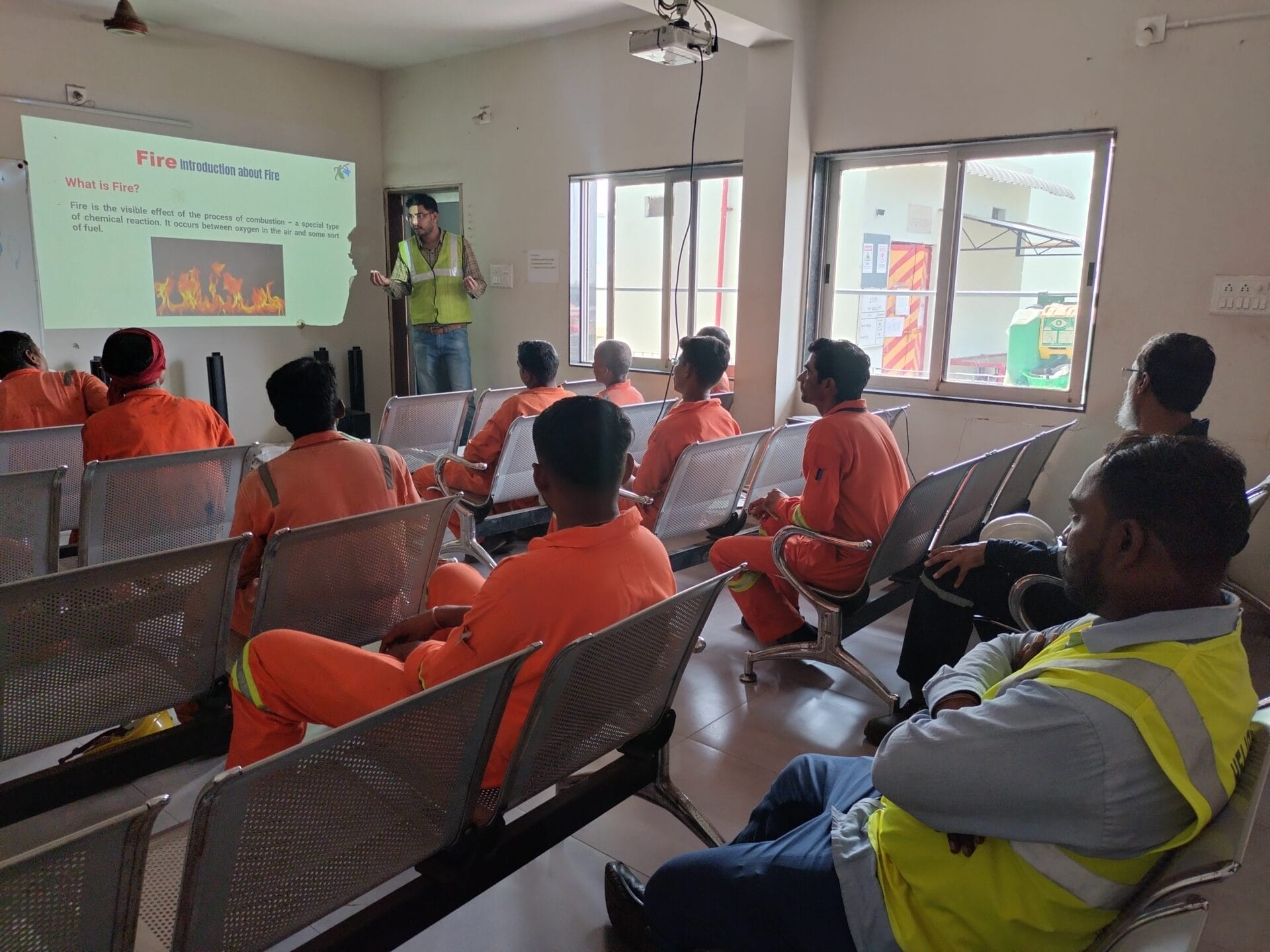 A group of people in orange uniforms sits in a classroom, listening to a presenter in a yellow vest give a Workplace Safety presentation with a slide about fire displayed on the screen.