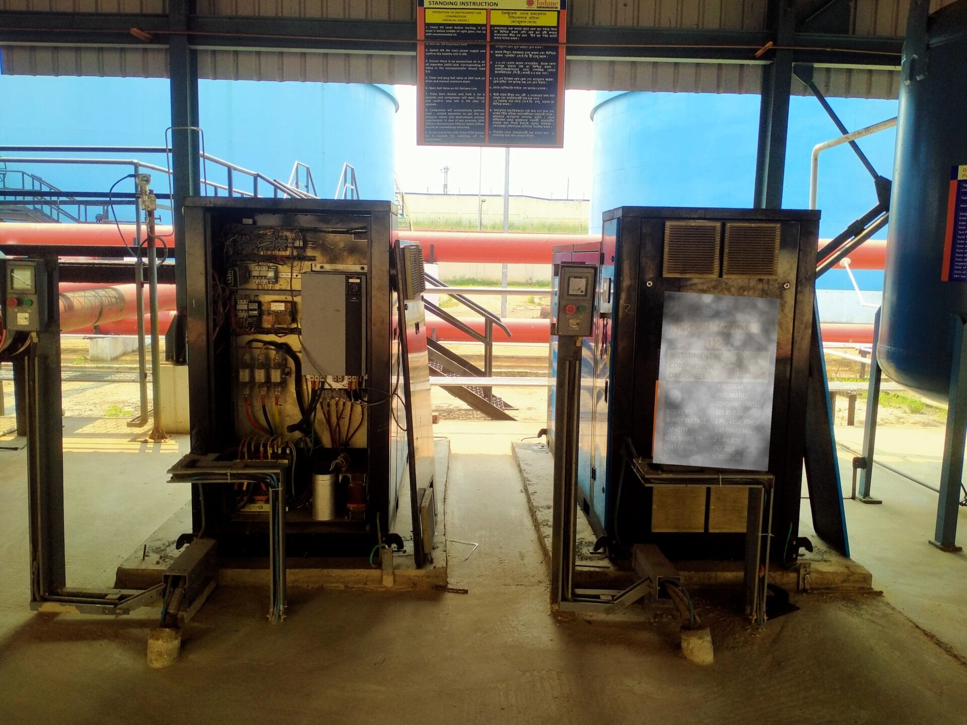 Two industrial control panels stand side by side in a facility, one open to reveal wiring and components vital for air quality analysis. Large blue storage tanks and red pipes appear in the background, highlighting the focus on environmental safety.
