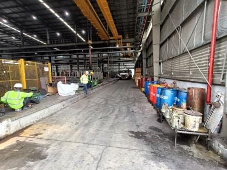 A spacious industrial warehouse in Kolhapur, Maharashtra, with two workers in safety gear, yellow fencing on the left, and stacks of colorful barrels on the right. Overhead cranes hint at ongoing Energy Efficiency improvements for greater cost savings.