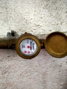 A brass water meter with an open lid is mounted on a rough, white wall. The dial displays numbers, the brand “Kranti,” and colored markings—ideal for water conservation efforts or industrial water use efficiency assessments.