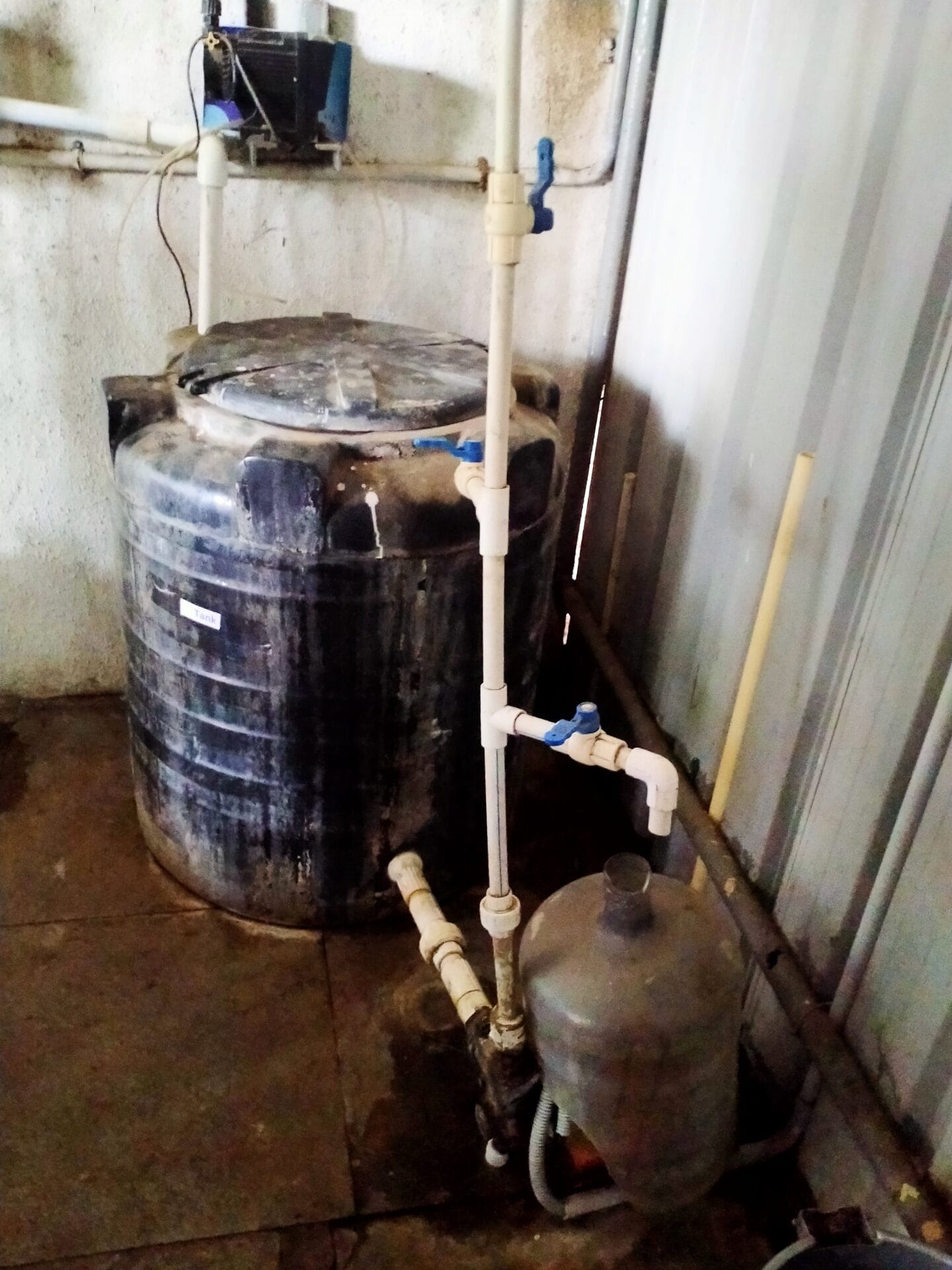 A large black plastic water tank connected to white PVC pipes and valves sits against a wall; a smaller container is linked beside it—part of an indoor utility area, ideal for conducting a water usage audit to improve efficiency.