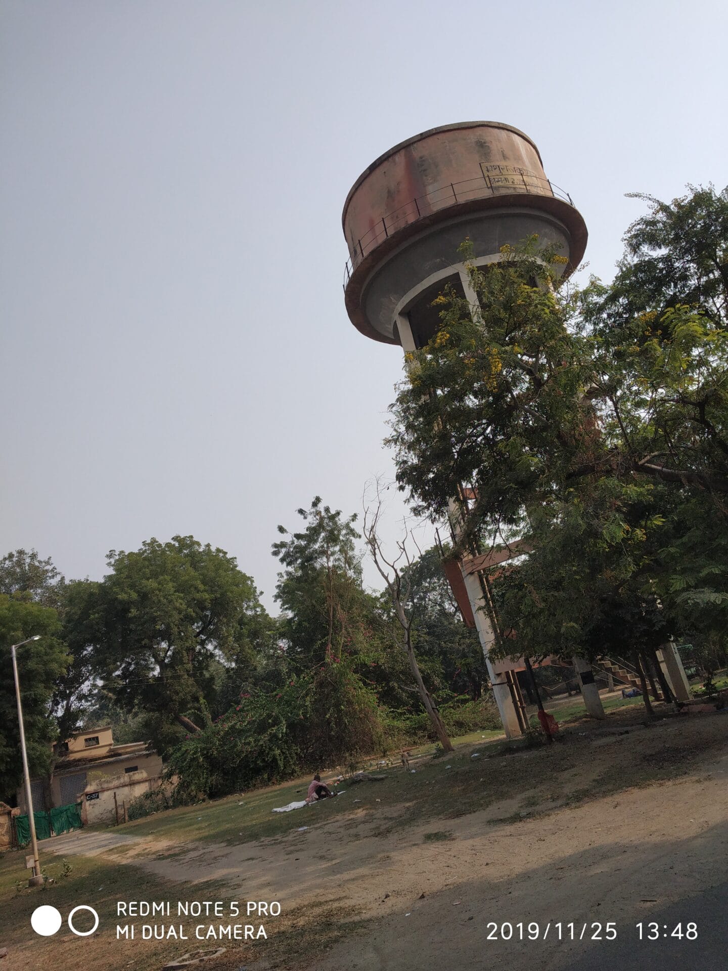 A tall, cylindrical water tower with a rusted tank stands near trees and buildings on a sunny day in Amritsar, Punjab. The bare ground and greenery reflect ongoing water conservation efforts. The photos timestamp is 2019/11/25.