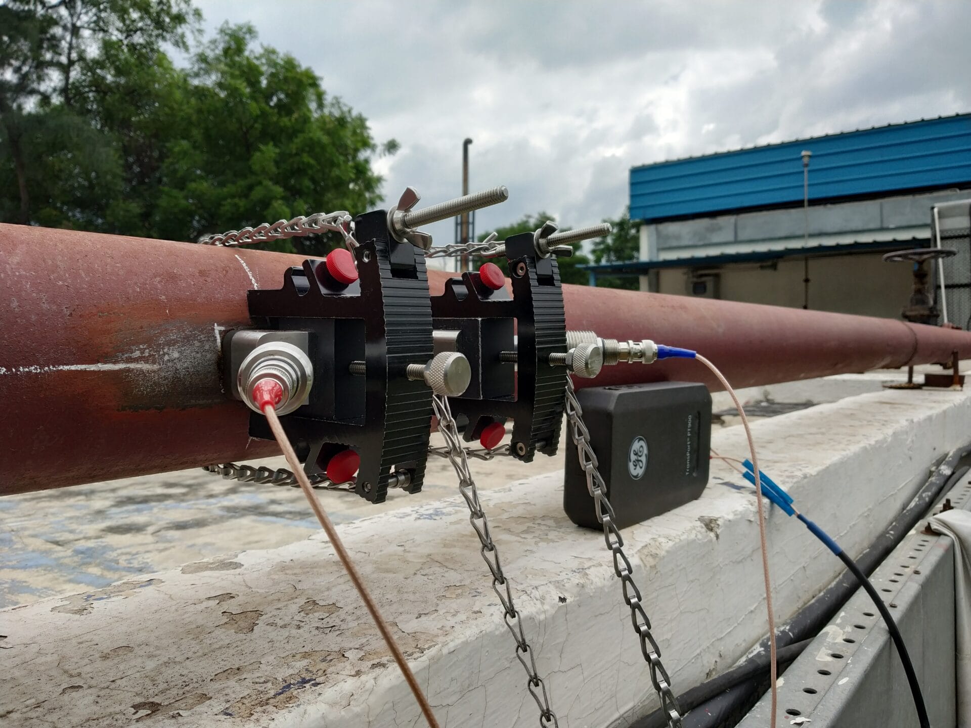 A close-up of a rusted metal pipe with sensor clamps and red adjustment knobs, used for Water Quality Analysis, connected to a device outdoors near a blue-roofed building and trees—possibly at a beverage plant in Noida.