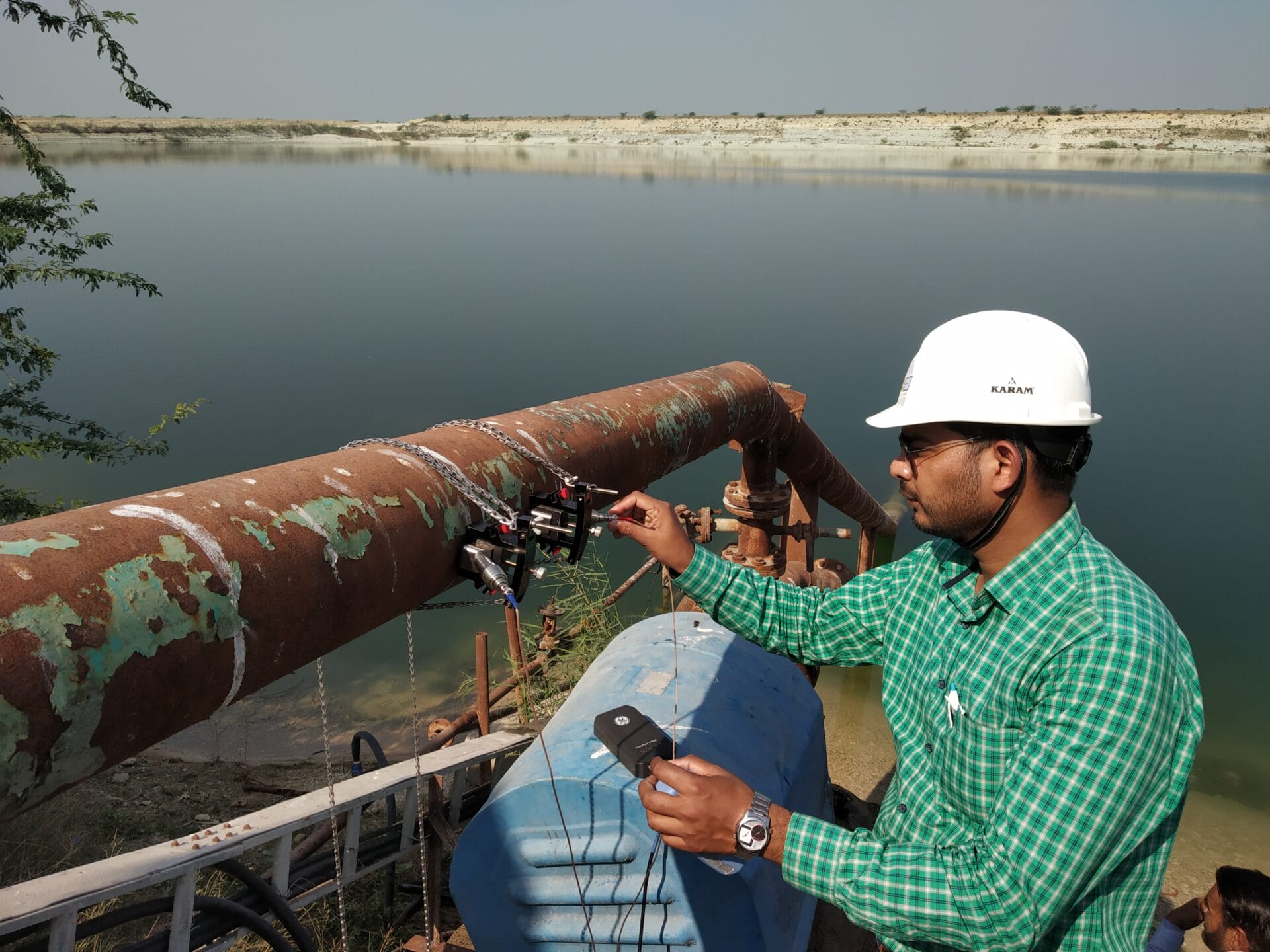 inspects a rusty pipeline using a handheld device near a large body of water, conducting a water efficiency audit with the rocky shoreline visible in the background.