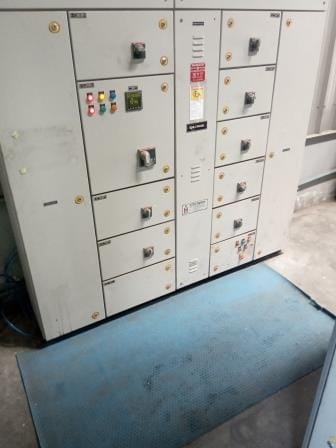An electrical control panel with multiple switches, indicator lights, buttons, and cabinet doors is positioned behind a blue anti-slip mat on a concrete floor, ensuring Electrical Safety and Compliance in workplace operations.
