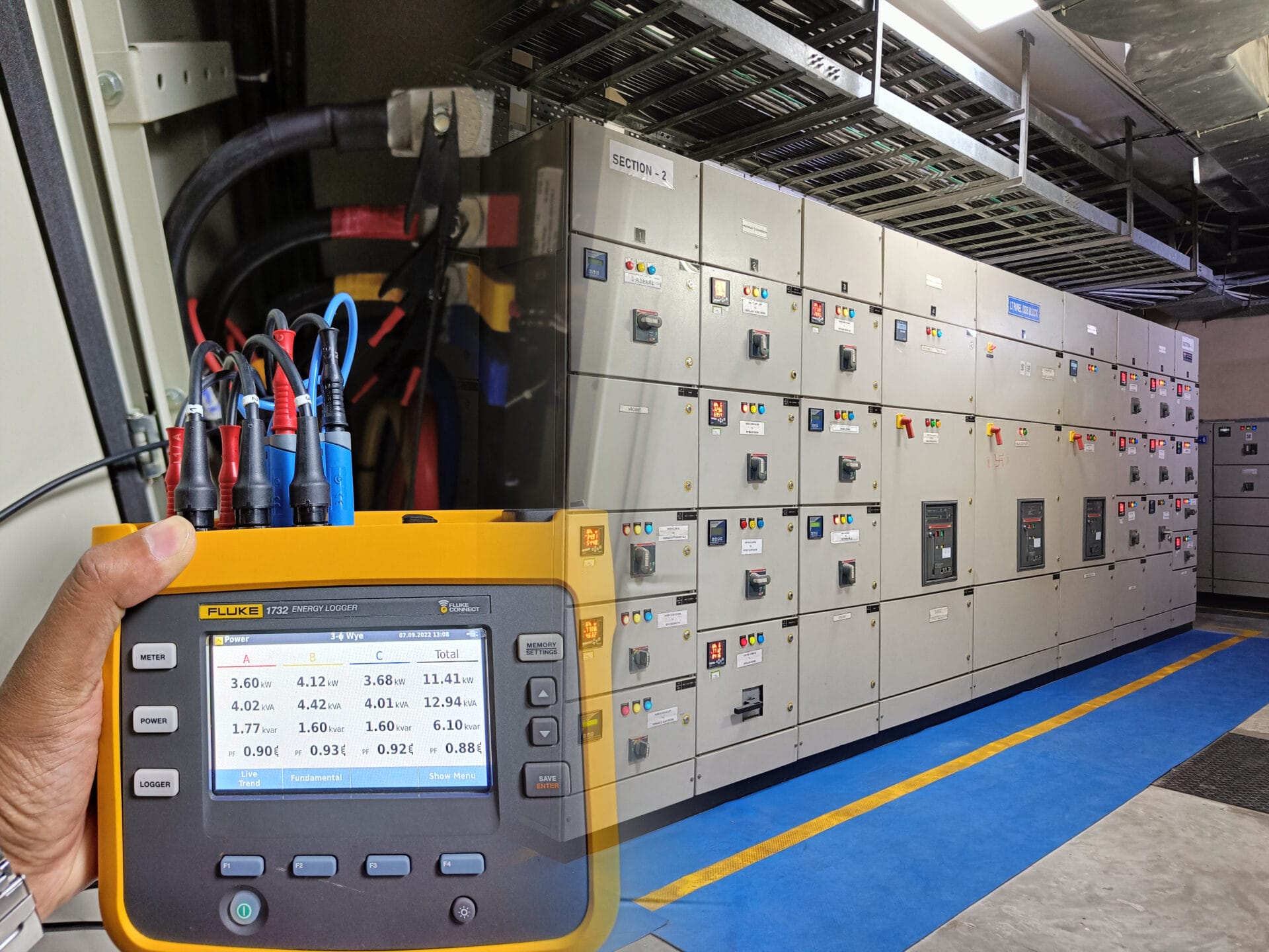 A hand holds a Fluke power meter displaying electrical readings in front of a large room filled with gray control panels, supporting preventive maintenance and energy efficiency across switchgear cabinets.