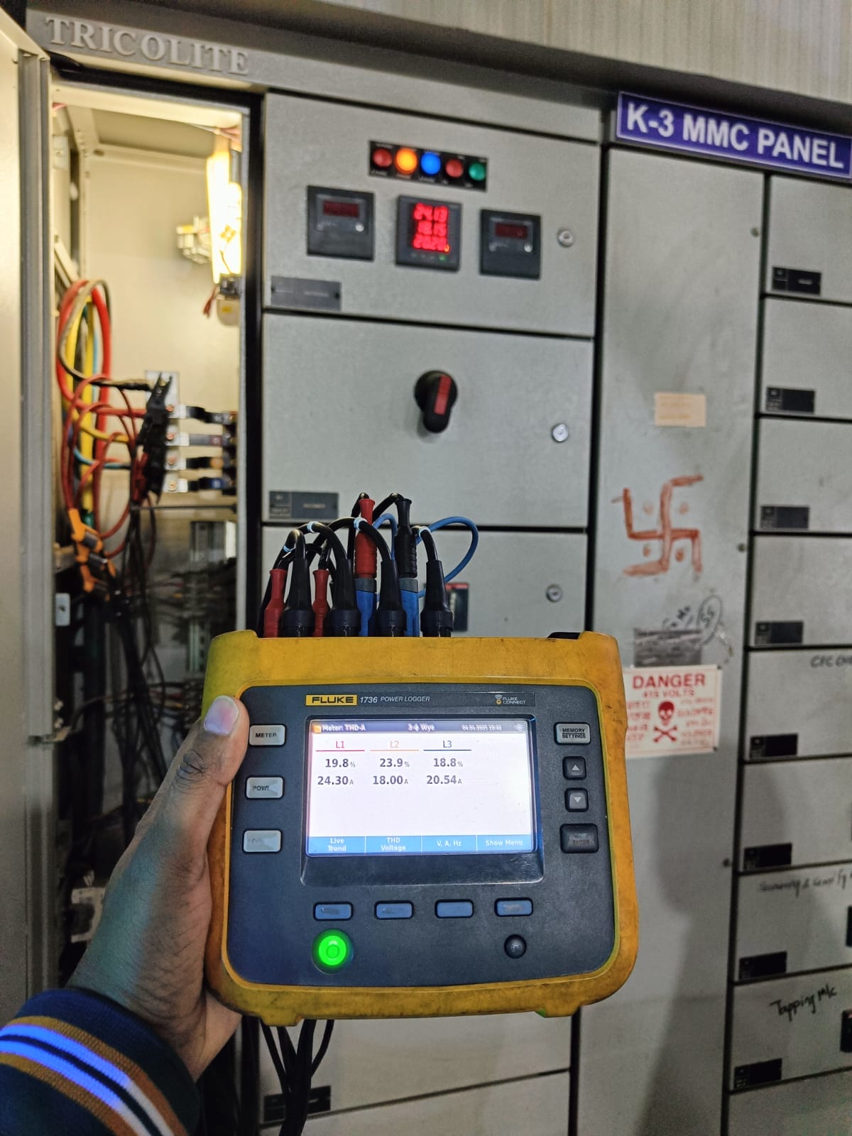 A hand holds a Fluke power quality analyzer with cables attached, in front of an open K-3 MMC PANEL. The display shows electrical readings, while warning labels are visible—ideal for use during an electrical thermography inspection.