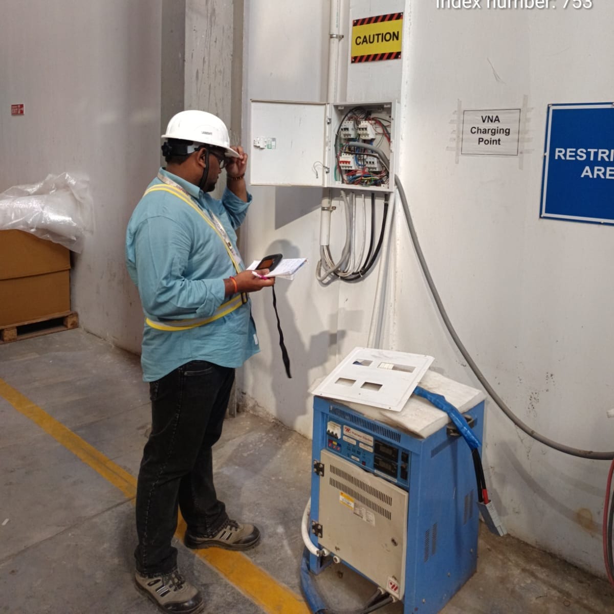 A worker in a safety helmet and vest inspects an open electrical panel labeled VNA Charging Point during an electrical safety inspection, holding a device and clipboard near a blue charging unit in an industrial setting.