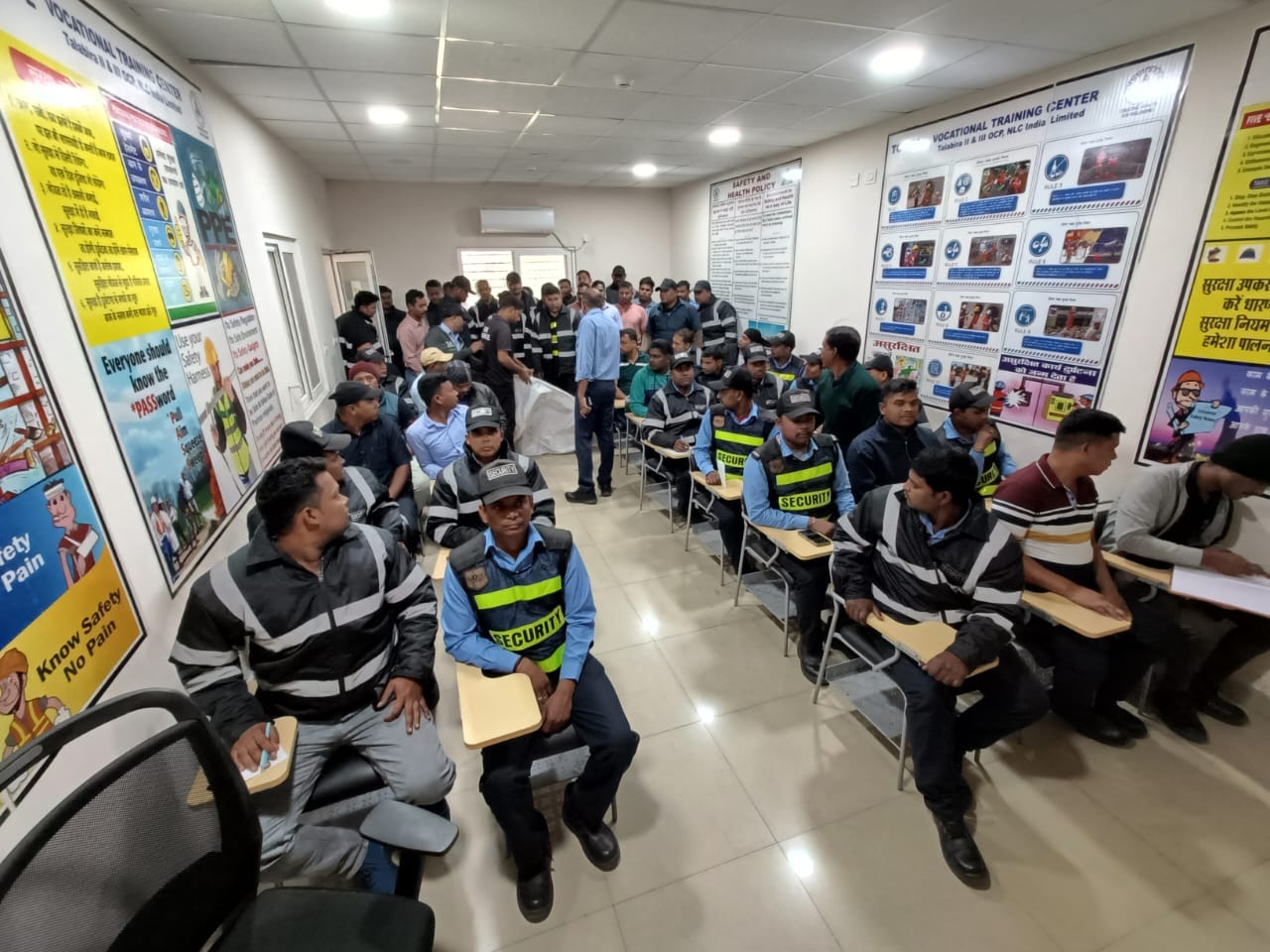 A classroom filled with adults, many in security uniforms, seated at desks. Instructors lead customised safety training as colorful workplace safety and emergency preparedness posters cover the walls.