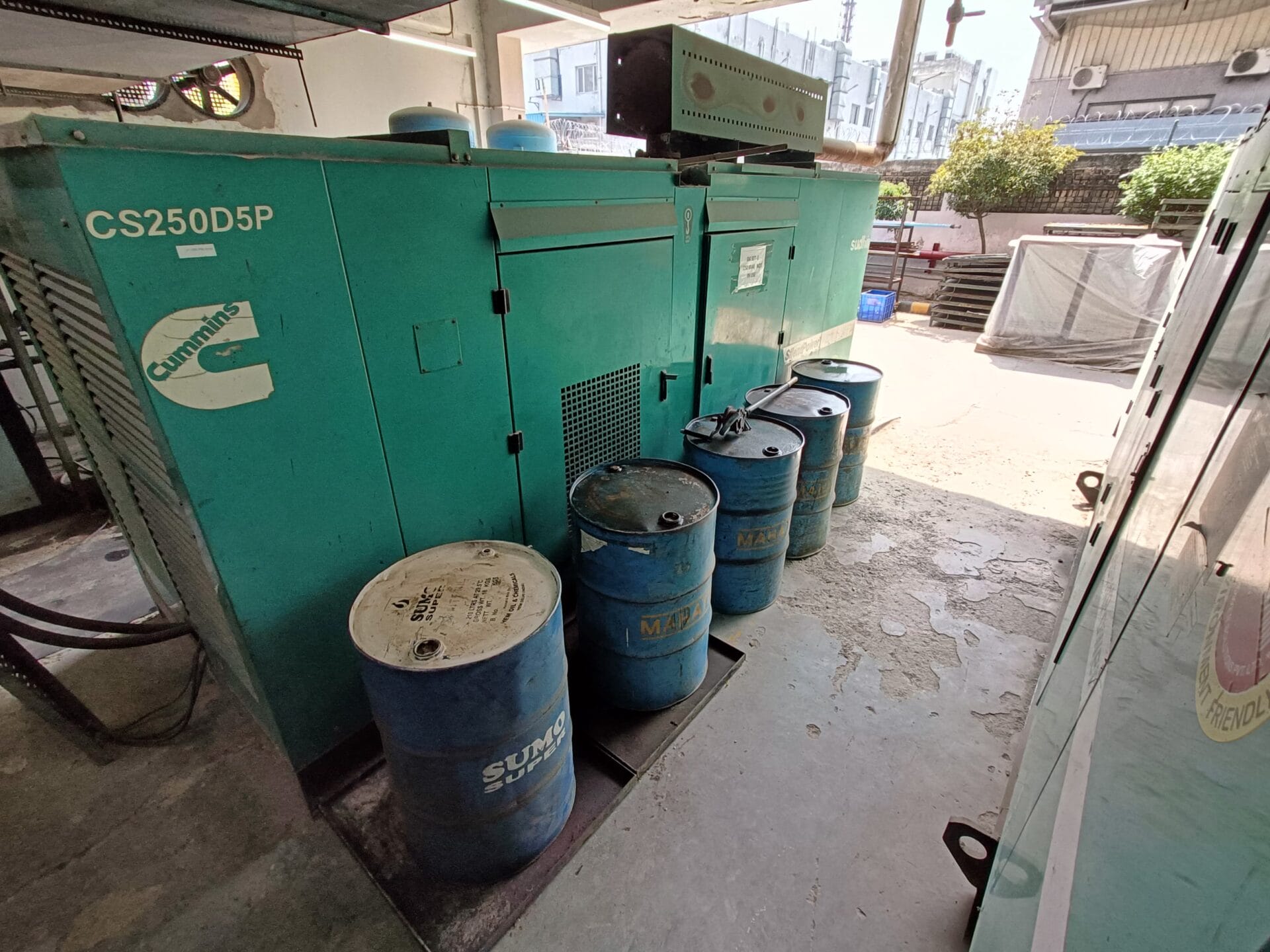 Several blue industrial barrels are placed beside large green Cummins generators outdoors, with buildings and air conditioning units visible in the background.