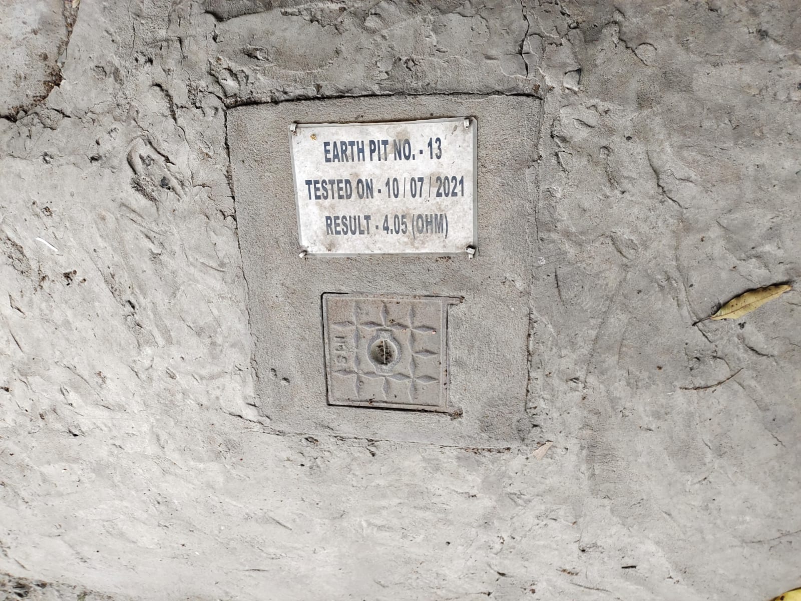 A metal plate labeled EARTH PIT NO.-13 is embedded in rough concrete, indicating an Earth Pit Testing on 10/07/2021 with a result of 4.05 ohm, ensuring electrical safety and compliance for the earthing system.