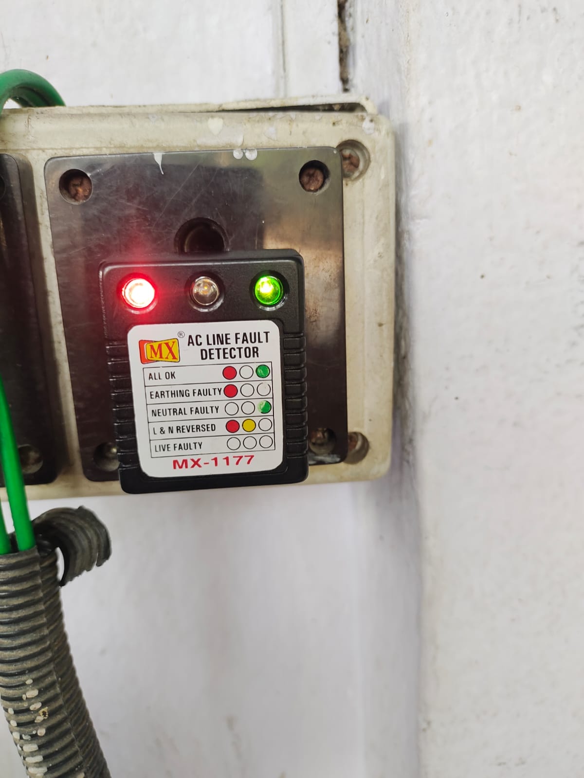 A close-up of an MX AC line fault detector (model MX-1177) plugged into a wall socket, showing one red and one green indicator light illuminated—ideal for use during an electrical safety audit. A green wire and conduit are visible on the left.