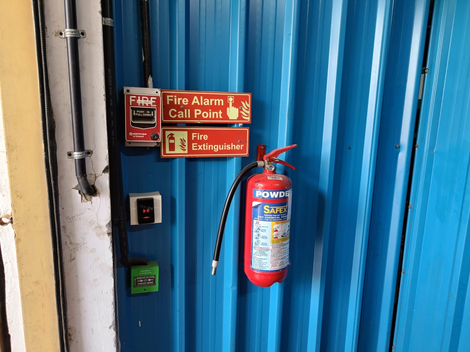 A blue metal wall with a mounted fire extinguisher beneath a red fire alarm call point sign—ideal for a fire safety inspection or fire risk assessment, featuring both a fire alarm button and a green emergency button nearby.