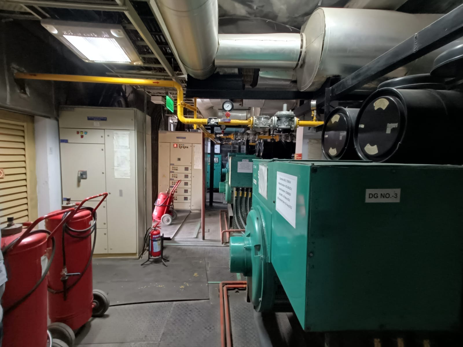 A room with industrial equipment, including large green machines labeled DG NO.3, red fire extinguishers, control panels, and metal pipes running along the ceiling—ideal for a workplace energy safety assessment or audit in a generator or machinery room.