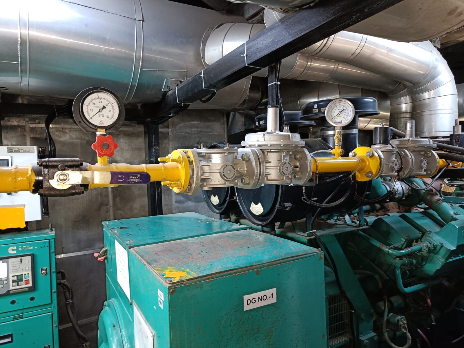 Yellow gas pipeline with pressure gauges, valves, and meters connected to industrial green machinery, likely in a mechanical or generator room with large metal ducting overhead—ideal for workplace energy safety and risk management practices.