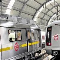 Two silver Metro Rail trains with yellow stripes and red logos are stopped at an indoor, arched-roof station platform in Delhi NCR, reflecting a high standard of public transport safety and compliance.