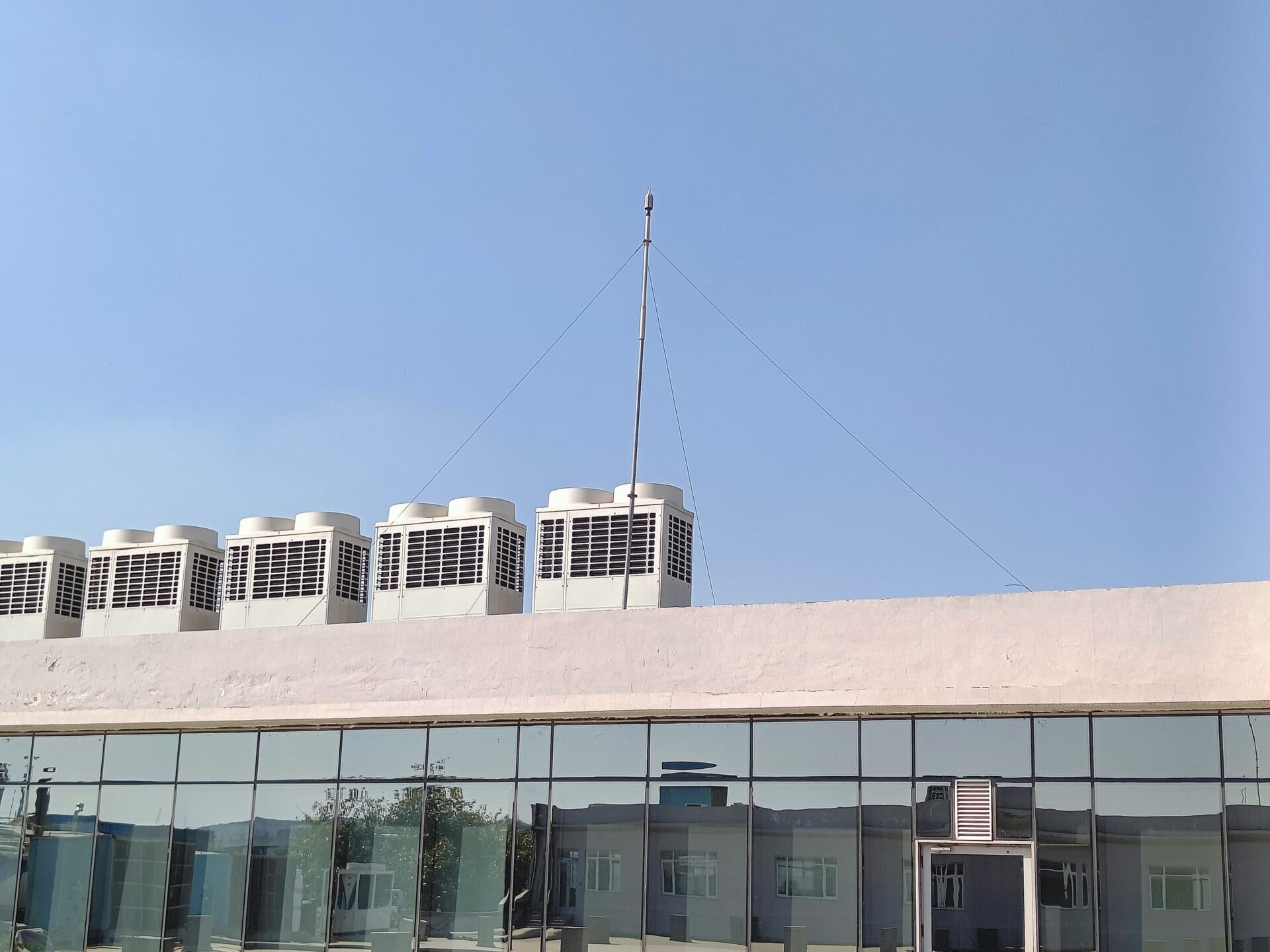 A row of air conditioning units sits on the flat white roof of a building with large reflective glass windows, ensuring optimal HVAC efficiency under a clear blue sky. An antenna, supported by guy wires, is mounted on the roof.