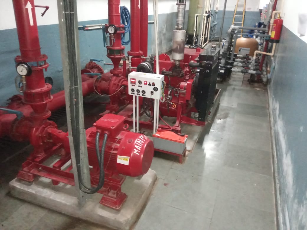 Red industrial fire pump system with pipes, gauges, a control panel, and motors installed indoors along a blue and white wall; valves and piping are visible in the background.
