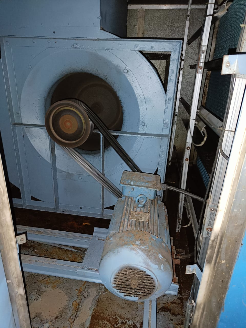 A large industrial fan with a visible motor and belt drive system, partially enclosed in a blue metal frame, operates in a machinery room—ideal for review during an industrial energy audit or compressed air system assessment. Fan blades and belts are in motion.