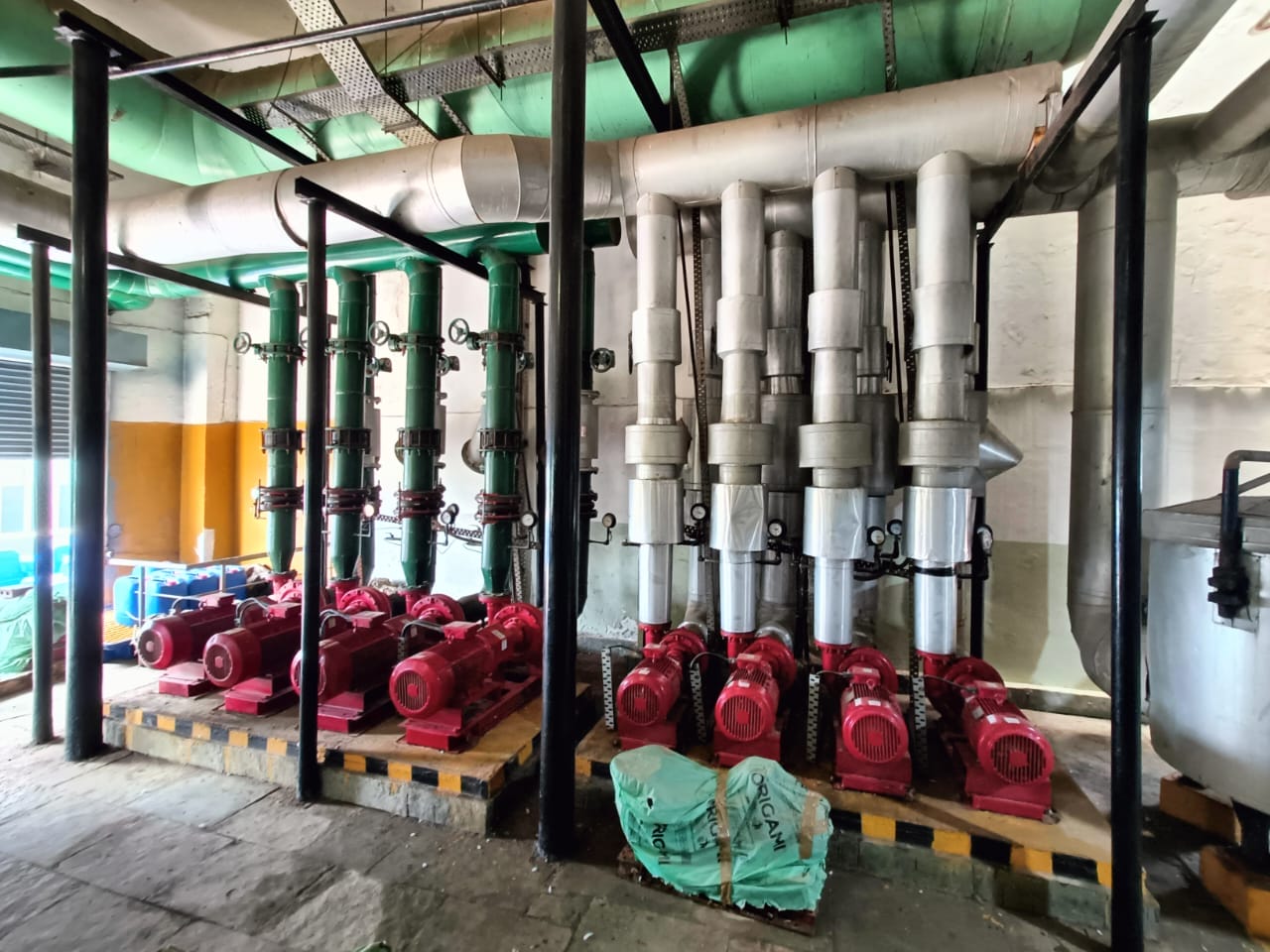 A row of industrial red pumps connected to vertical metal pipes and valves in a utility room, featuring green overhead ducts and a concrete floor. Equipment and materials are scattered nearby.