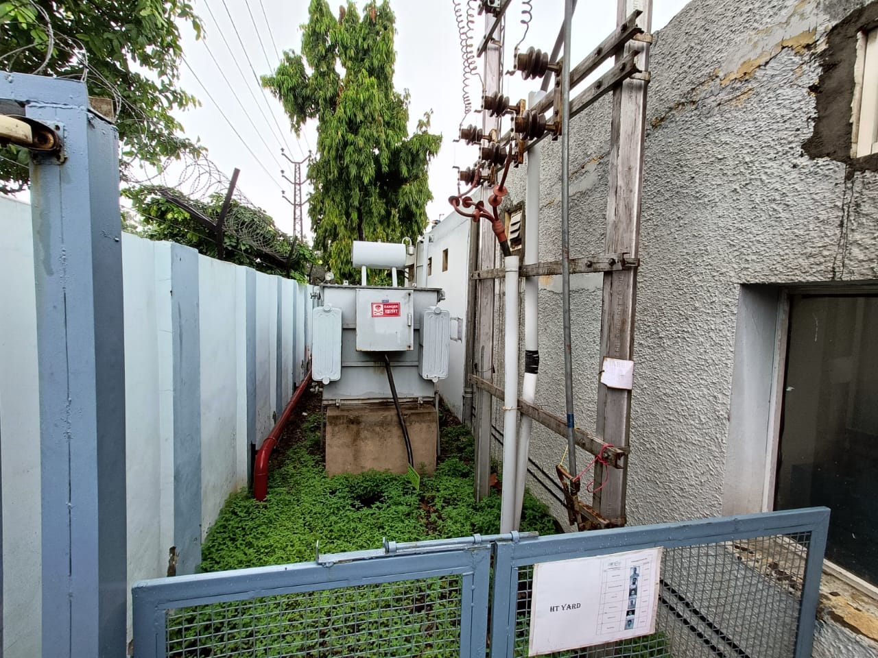 A large electrical transformer is installed outdoors in a fenced enclosure, surrounded by green ground cover and a grey wall. Overhead power lines and equipment are visible above—an ideal setting for an industrial safety assessment.