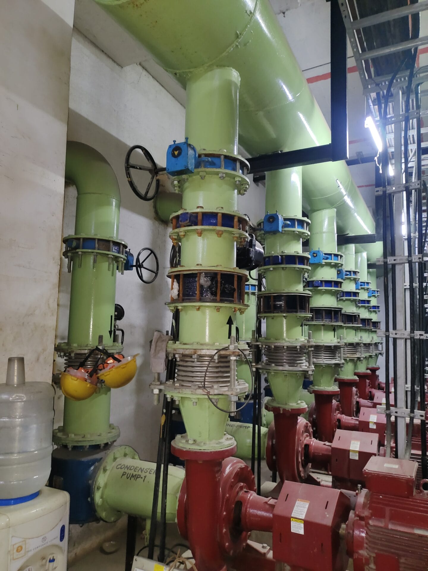 Large green industrial pipes with blue valves and red pumps run vertically and horizontally inside a utility room in Hyderabad, Telangana. Nearby, a water dispenser, safety equipment, and an electrical panel assessment area are visible.
