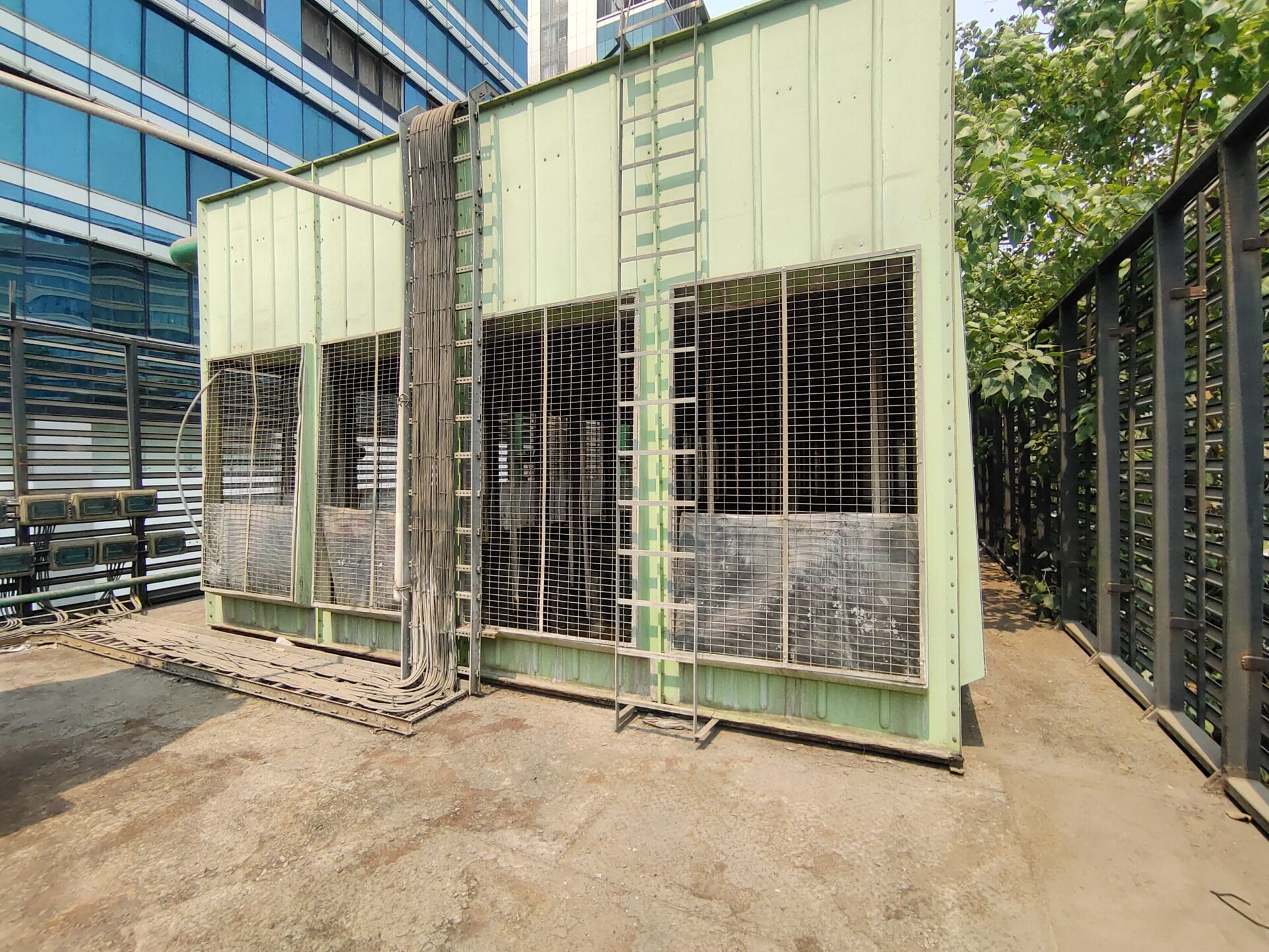 A large green industrial structure with metal mesh panels, ladders, and pipes stands outdoors on a concrete surface near a modern glass building and trees, likely serving as part of an air filtration system study for data center air quality in Hyderabad Telangana.